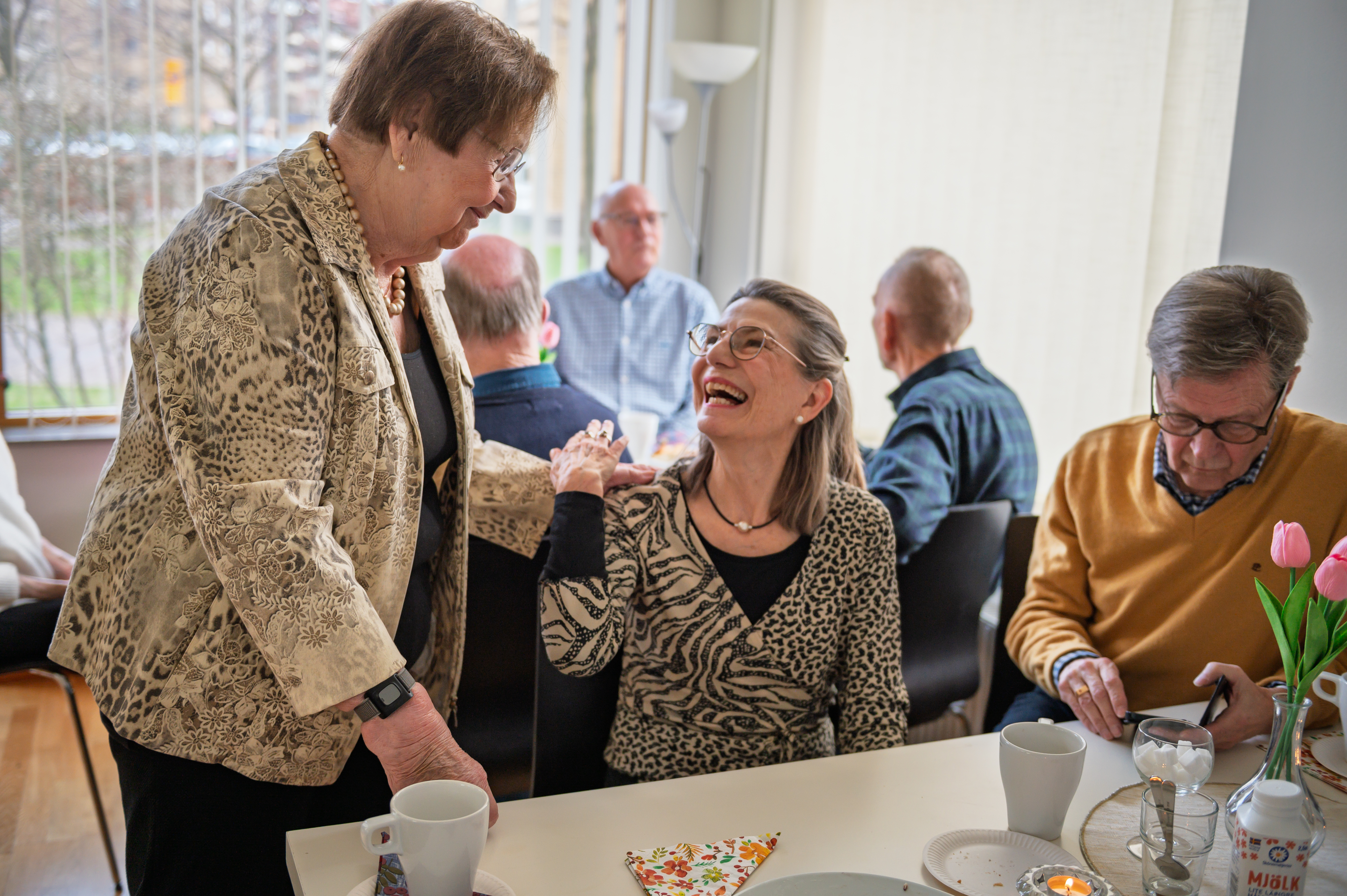 Två personer hälsar på varandra i ett fikarum där andra personer sitter runtomkring.
